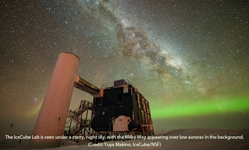 The IceCube Lab is seen under a starry, night sky, with the Milky Way appearing over low auroras in the background.